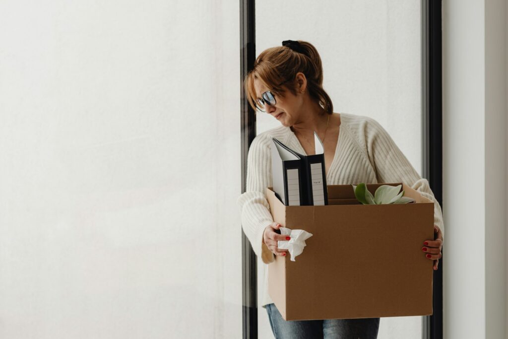 A woman holding a box of her personal effects from work, looking sad, representing employee termination