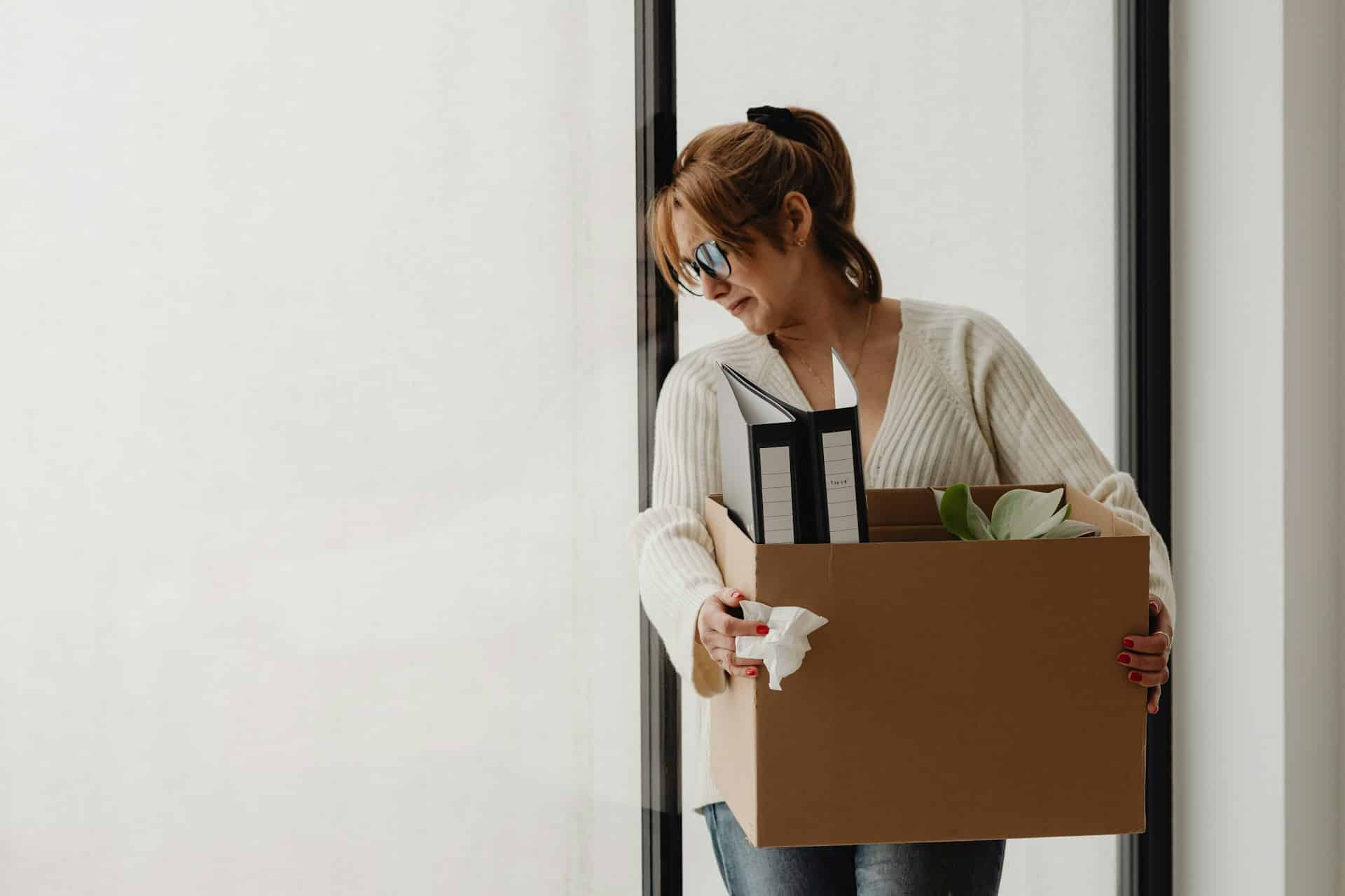 A woman holding a box of her personal effects from work, looking sad, representing employee termination