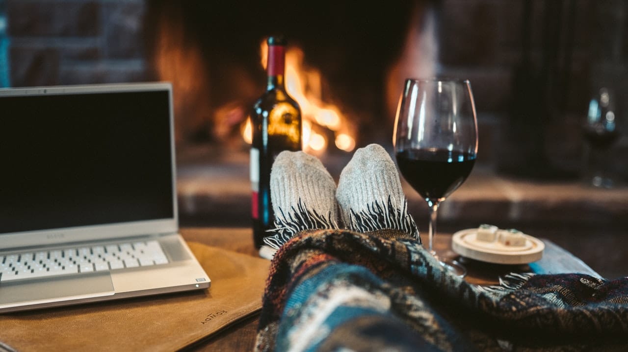 A person's feet up on a coffee table by a fire with a glass of wine and a laptop, representing remote holiday work parties during COVID-19