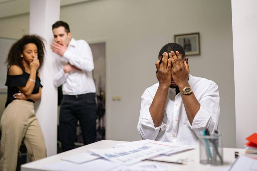 A man with his face in his hands at his desk while coworkers whisper about him in the background, representing poisoned work environments and constructive dismissal