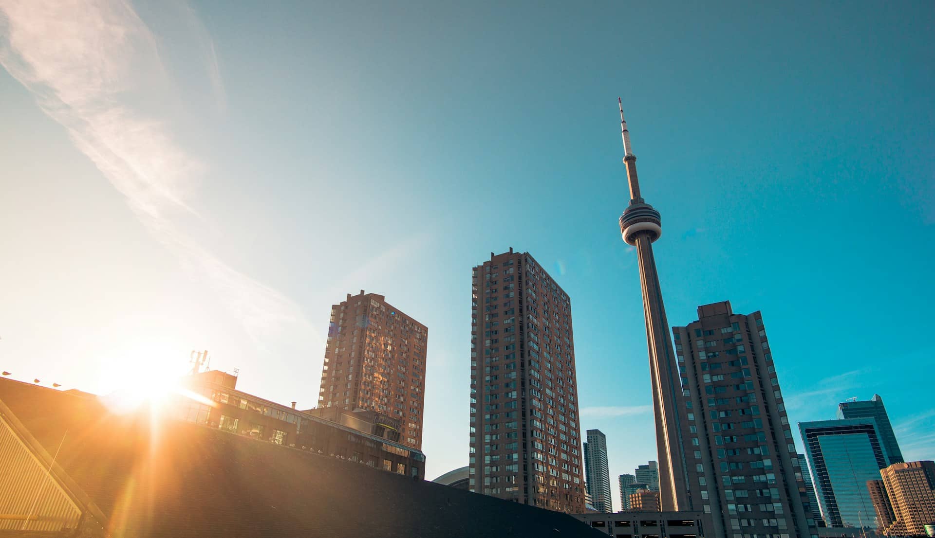 A low-angle shot of the CN Tower and Toronto office buildings rising up against the clear blue sky, with the morning sun glaring in the left corner, representing Best Law Firms Canada.