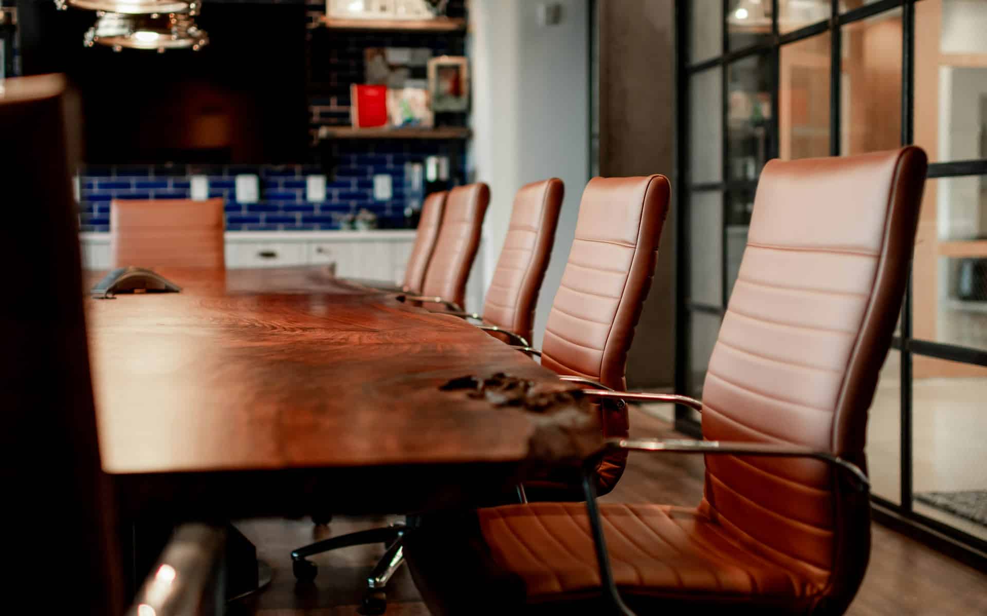 The right corner of a wooden meeting table with empty brown leather chairs pushed in, set in a modern boardroom, representing first-time collective bargaining.