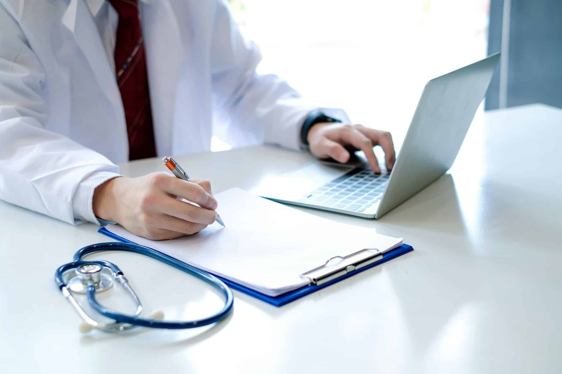 A doctor, shown from the shoulder down, sits at a white desk, while typing on a laptop with one hand and writing on a clipboard with the other. His stethoscope is curled next to the clipboard. This image represents sick notes required by employers for an employee's absence.