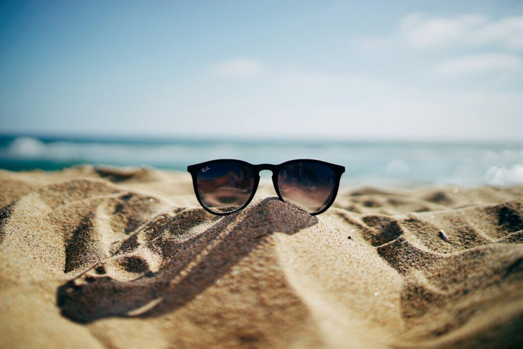 Sunglasses perched on a hill of sand on a beach with a blurred ocean stretching out under a soft blue sky, representing unlimited vacation policies.