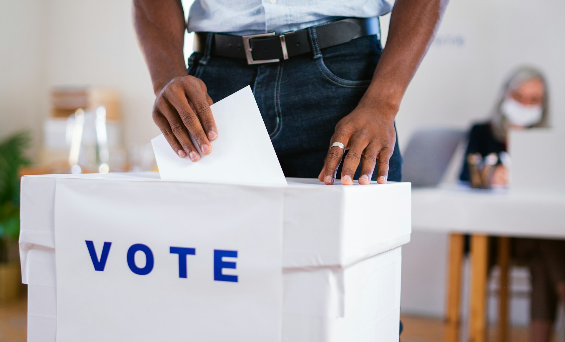A person, shown from the waist down, stands behind a white box with the word 'Vote' in blue across the front, slipping a piece of paper with their vote into it, representing union vote challenges.